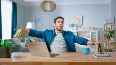 A happy investor sits at his desk in front of his laptop and does the mexican wave with his arms to celebrate the returns from his ASX dividend shares