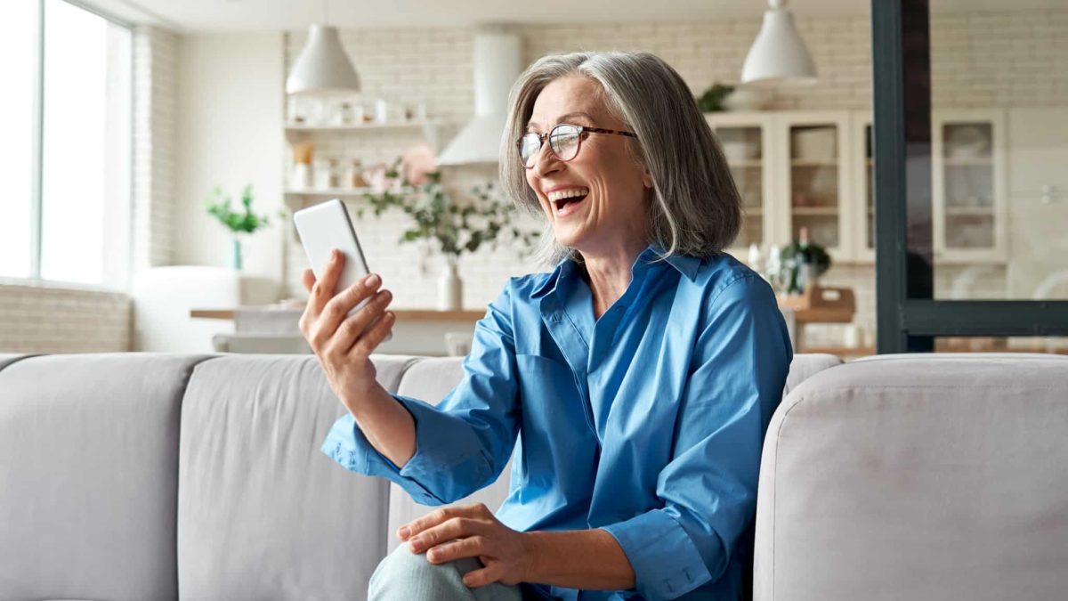 A sophisticated older lady with shoulder-length grey hair and glasses sits on her couch laughing while looking at her phone