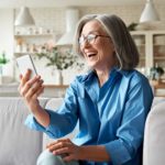 A sophisticated older lady with shoulder-length grey hair and glasses sits on her couch laughing while looking at her phone