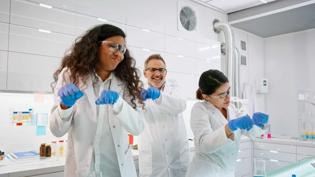 Three Archer Materials scientists wearing white coats and blue gloves dance together in their lab after making a discovery