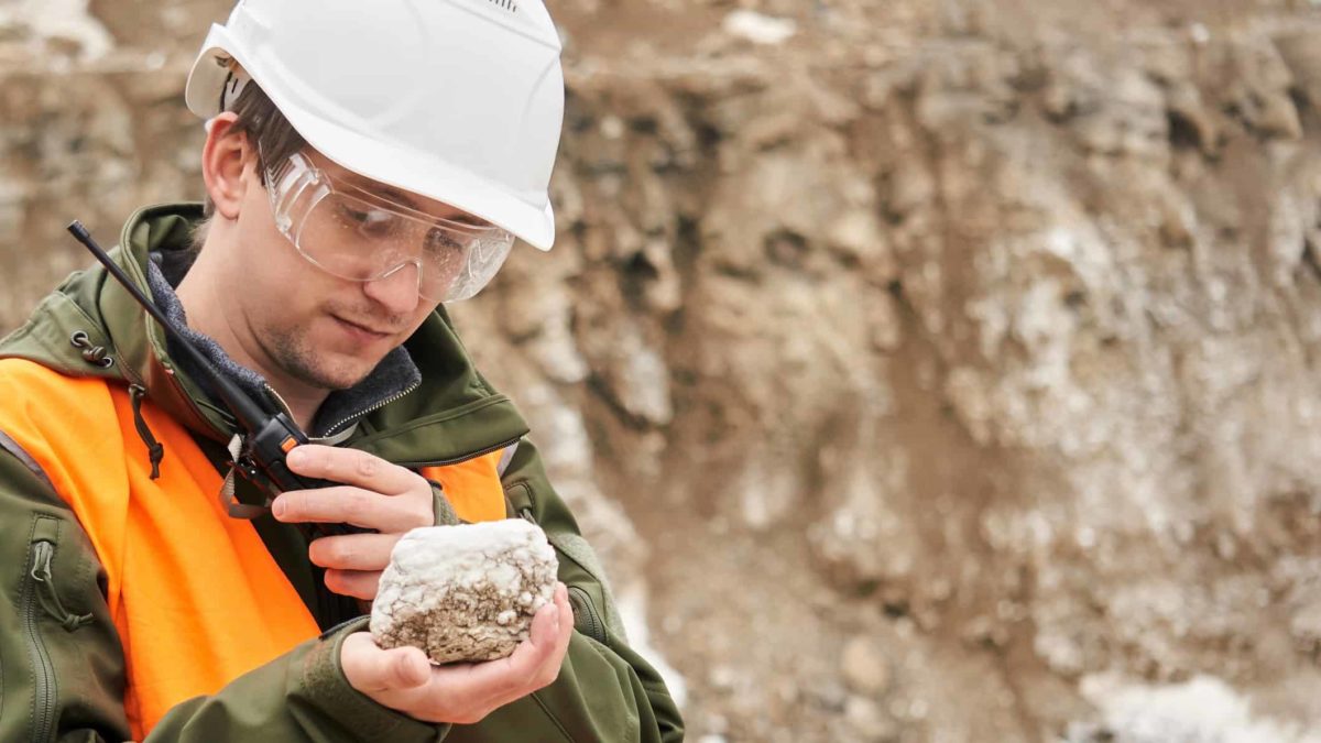 A male geologist wearing a white hardhat and orange high vis vest talks on a walkie-talkie while staring at a rock showing mineral deposits