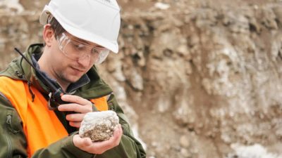 A male geologist wearing a white hardhat and orange high vis vest talks on a walkie-talkie while staring at a rock showing mineral deposits