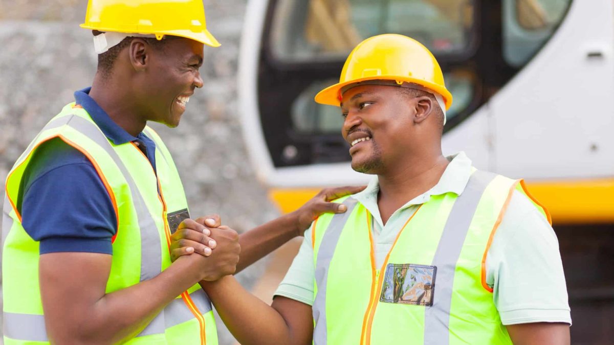 Two excited mining workers in yellow high vis vests and hardhats shake hands to congratulate each other on a mineral discovery