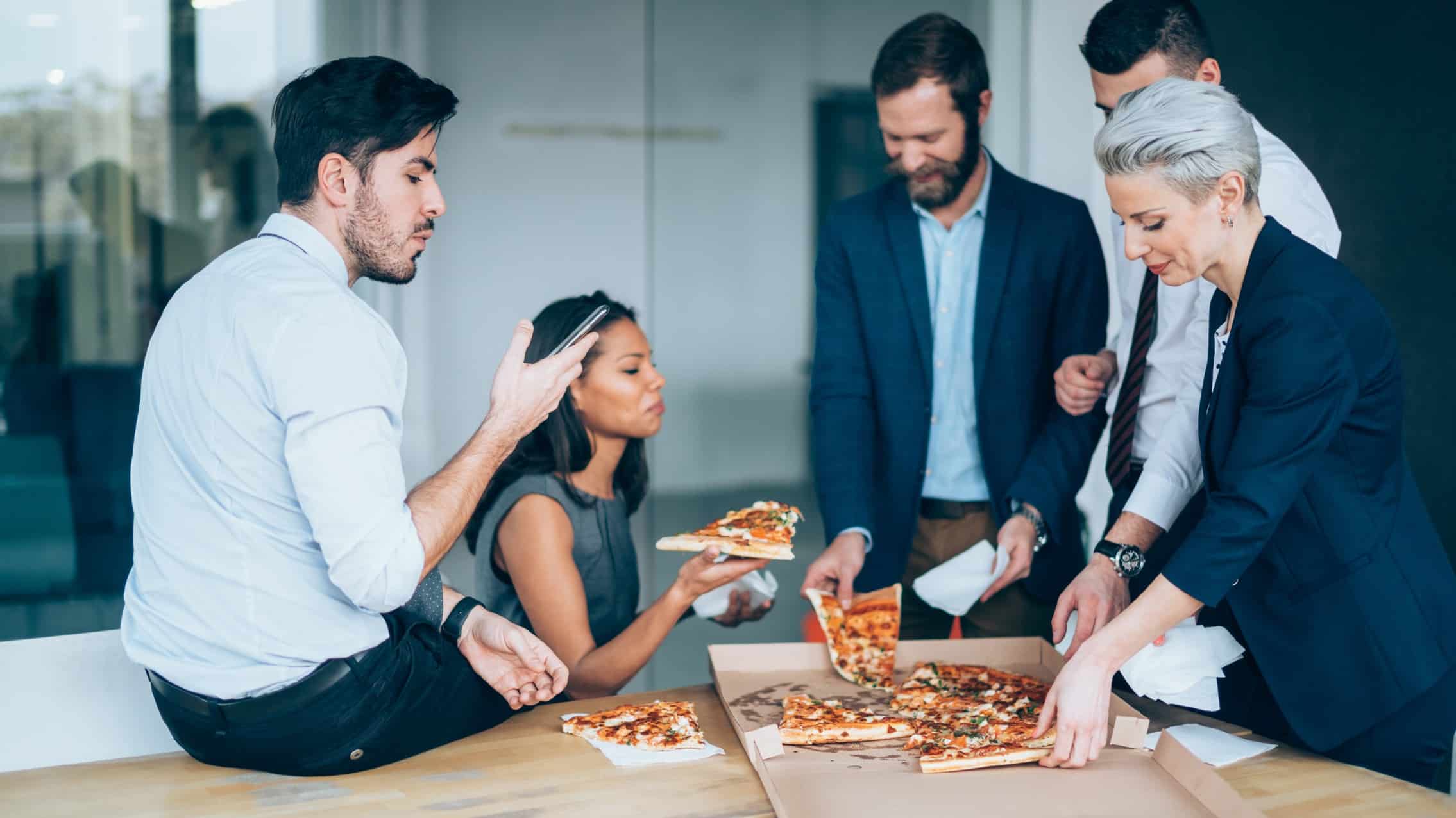 A team in a corporate office shares a pizza while standing around a table chatting about the Domino's share price and Pizza Hut's threat to the business