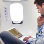 Man sitting in a plane seat works on his laptop.