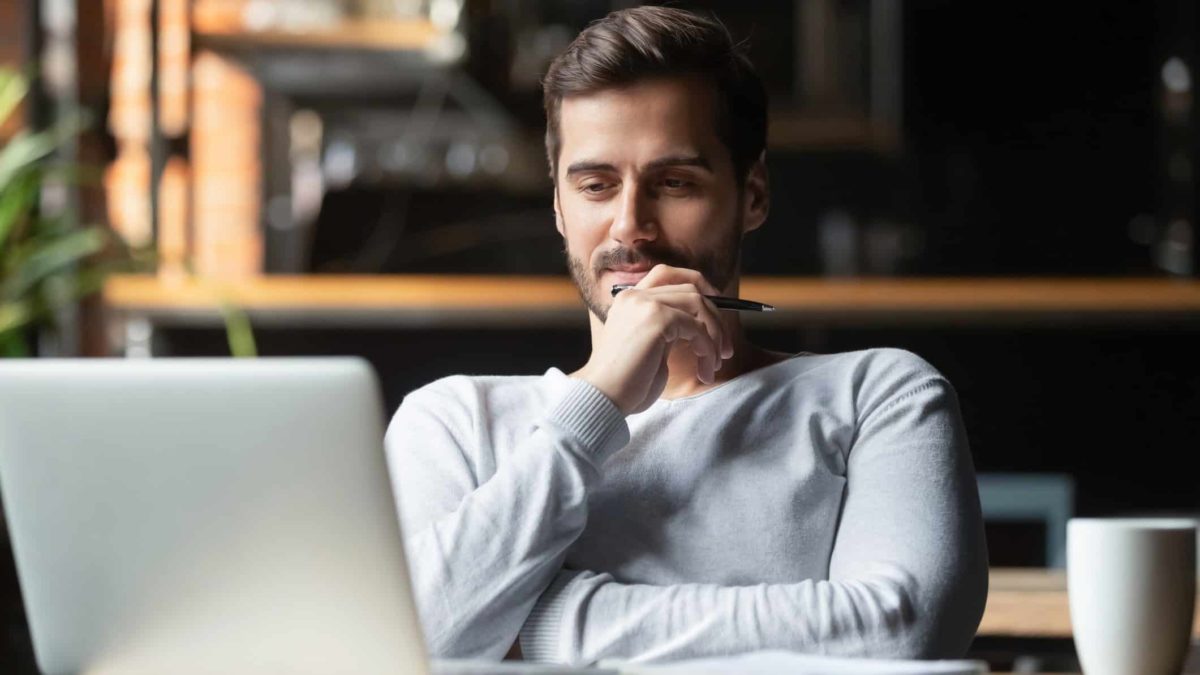 A male investor sits at his desk looking at his laptop screen holding his hand to his chin pondering whether to buy Macquarie shares