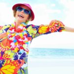 A woman wearing a bright multi-coloured dress, blue sunglasses and hat stands on a beach laughing with her arms outstretched enjoying herself