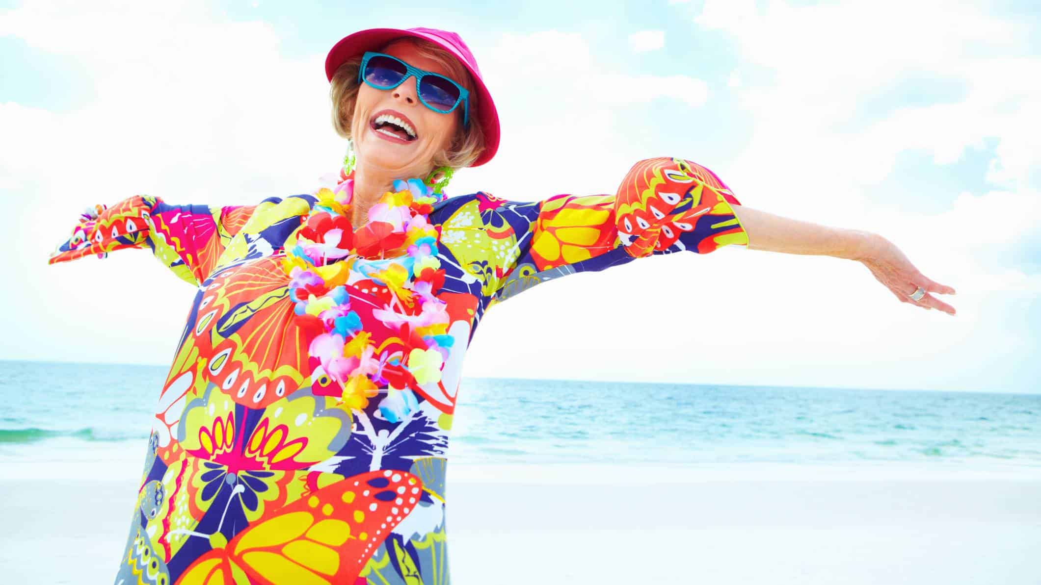 A woman wearing a bright multi-coloured dress, blue sunglasses, and hat stands on a beach laughing with her arms outstretched enjoying herself.