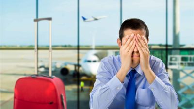 A man with a suitcase puts his head in his hands while sitting in front of an airport window.