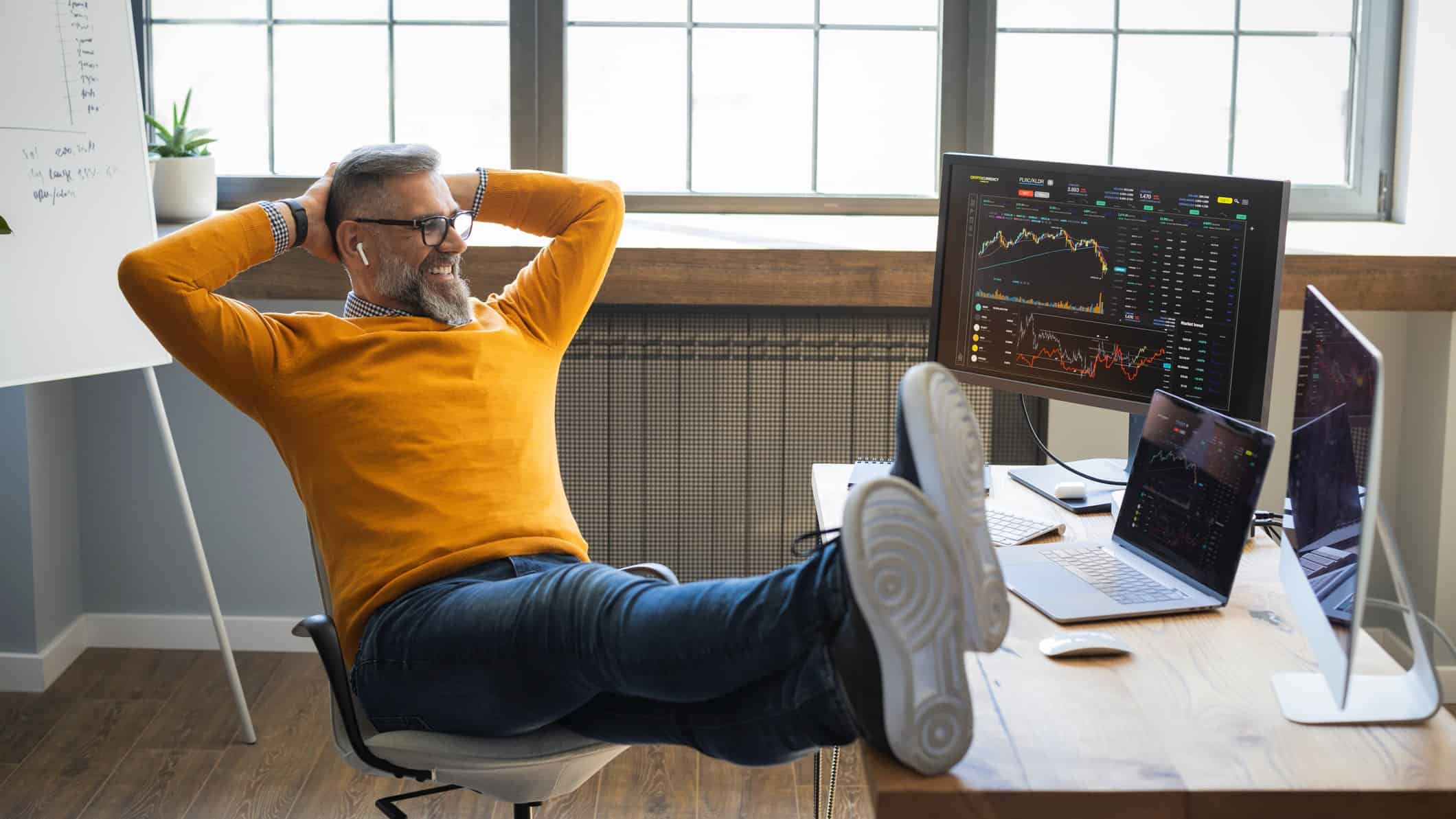 A man wearing glasses sits back in his desk chair with his hands behind his head staring smiling at his computer screens as the ASX share prices keep rising