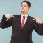 A smug investment manager in a suit and tie points to himself with both hands, feeling proud.