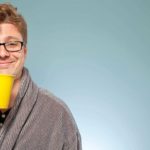 An investor wearing a dressing gown and holding a cup of coffee in a yellow mug gives a satisfied smile.
