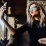 A smug executive woman wearing glasses and red lipstick blows a kiss to herself as she takes a selfie.