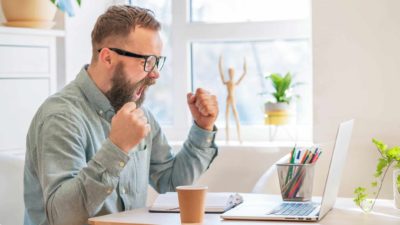 A young man wearing glasses and a denim shirt sits at his desk and raises his fists and screams with delight.