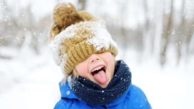 A young boy enjoys the snow with his beanie pulled down over his eyes as he sticks his tongue out to catch snowflakes