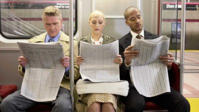 Two men and woman sitting in subway train side by side, reading newspaper