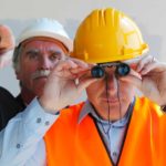 Man in yellow hard hat looks through binoculars as man in white hard hat stands behind him and points.