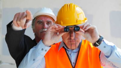 Man in yellow hard hat looks through binoculars as man in white hard hat stands behind him and points.