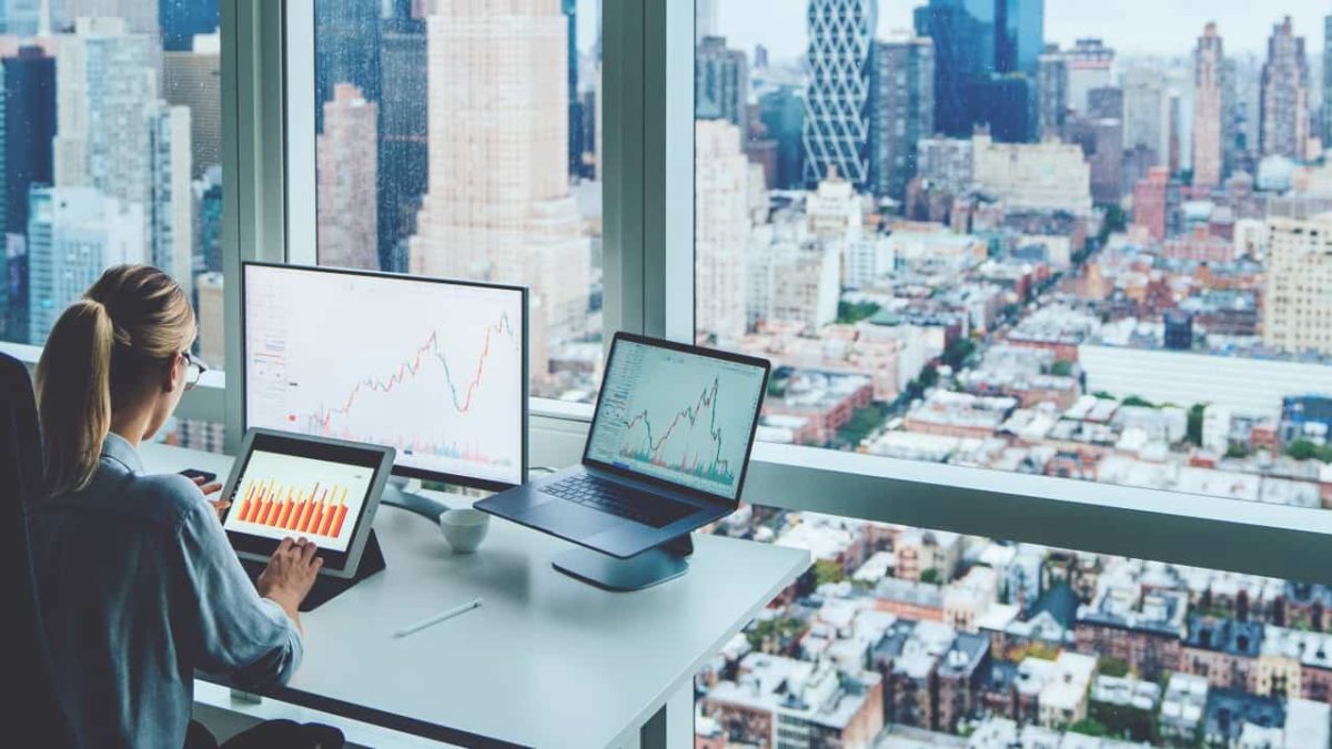 A female stockbroker reviews share price performance in her office with the city shown in the background through her windows