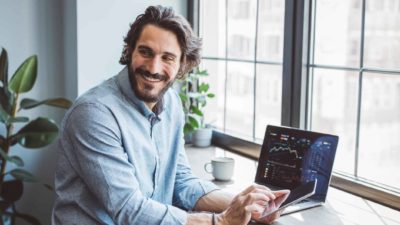 A happy male investor turns around on his chair to look at a friend while a laptop runs on his desk showing share price movements