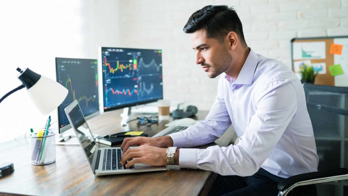 A male sharemarket analyst sits at his desk looking intently at his laptop with two other monitors next to him showing stock price movements