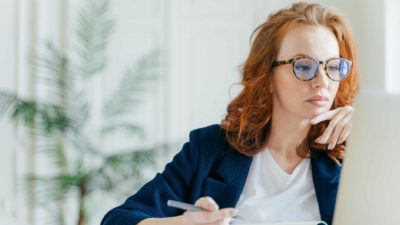 A female sharemarket analyst with red hair and wearing glasses looks at her computer screen watching share price movements.