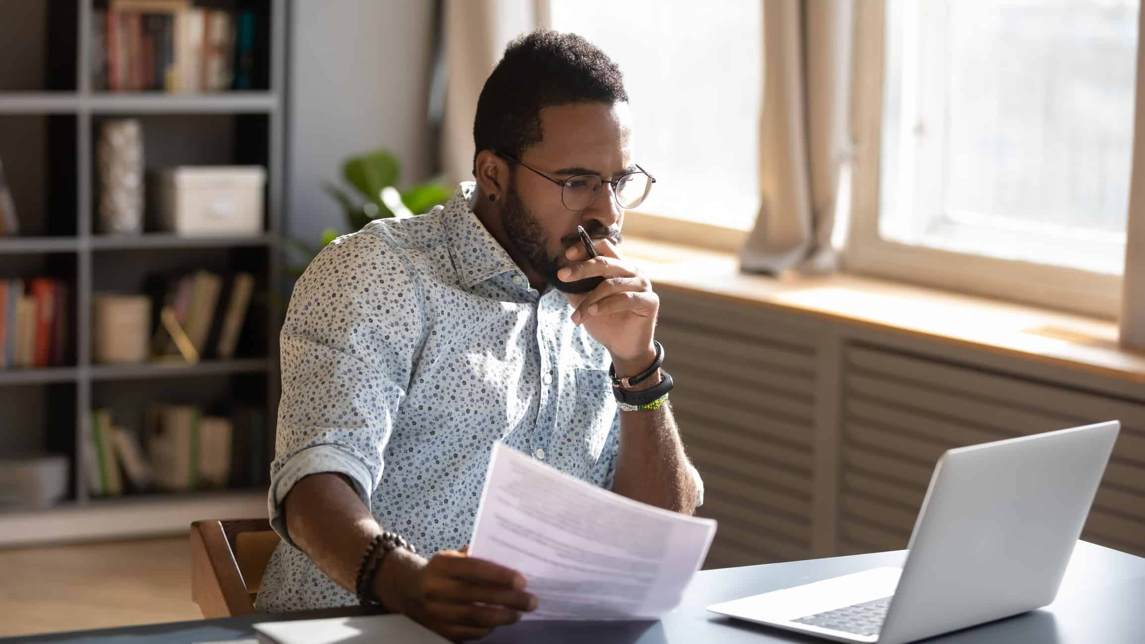 A male investor sits at his desk pondering at his laptop screen with a piece of paper in his hand.