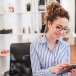 A young female investor sits in her home office looking at her ipad and smiling as she sees the QBE share price rising