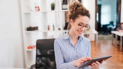 A young female investor sits in her home office looking at her ipad and smiling as she sees the QBE share price rising