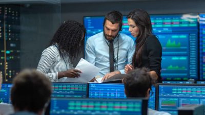 A trio of ASX shares analysts huddle together in an office with computer screens all around them showing share price movements