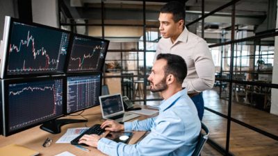 Two male ASX 200 analysts stand in an office looking at various computer screens showing share prices