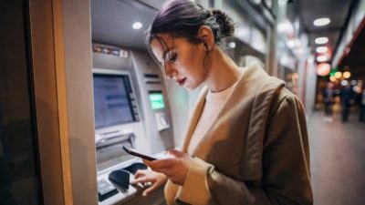 a woman looks at her phone while standing at an ATM machine in a night-time lit urban landscape.