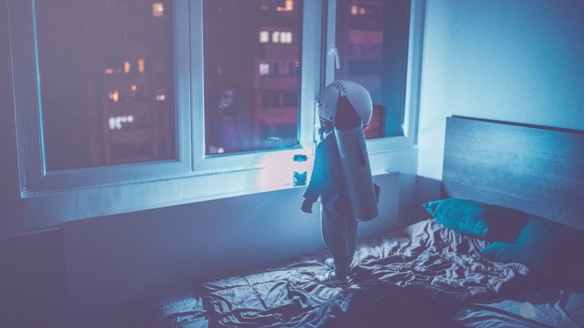 A boy stands on his bed at night looking at the city buildings outside, with a rocket on his back, ready to fly to the stars.