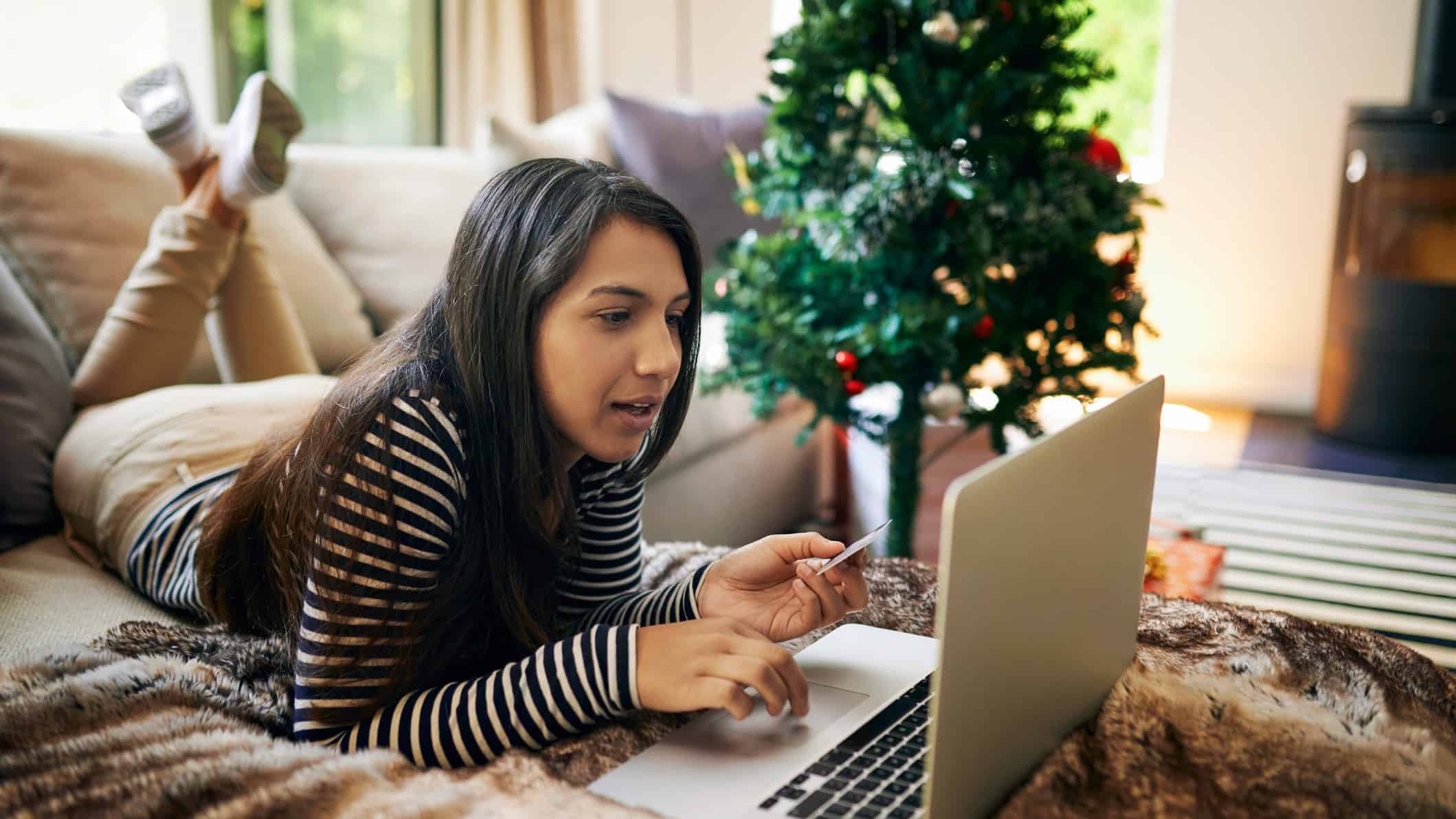 A young woman does her Christmas shopping online in her lounge room at home with a Christmas tree in the background.
