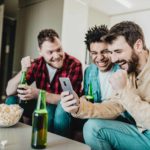A group of three young men sit on a sofa in a home environment with a bowl of popcorn and beer bottles in front of them cheering on one of their teams on a phone.