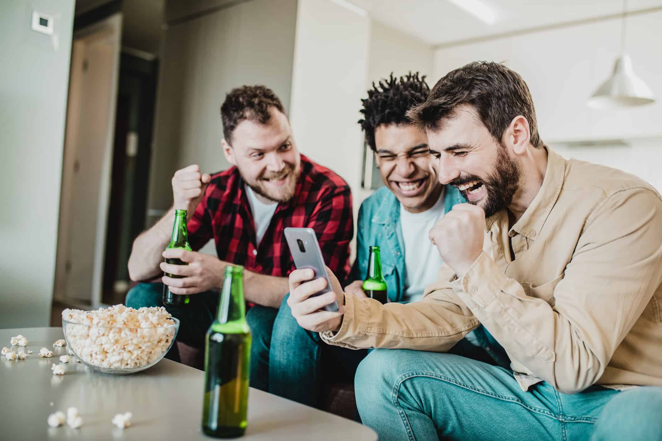 a group of three young men sit on a sofa in a home environment with a bowl of popcorn and beer bottls in front of them cheering on one of their group as he looks excitedly at his phone as though he's just had some success on an online gambling app.