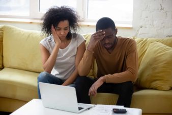 a young couple sit on their sofa at home looking distraught and downcast while sitting at an open laptop computer. The man has his head in his hand while tthe woman holds her hand to her face.