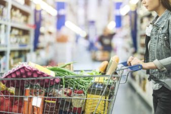 a woman stands with a full grocery trolley at the top of a supermarket aisle.