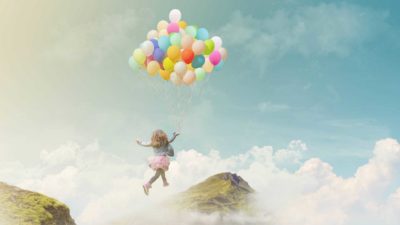 A girl holding a big bunch of balloons flies high through the clouds above the mountain tops.