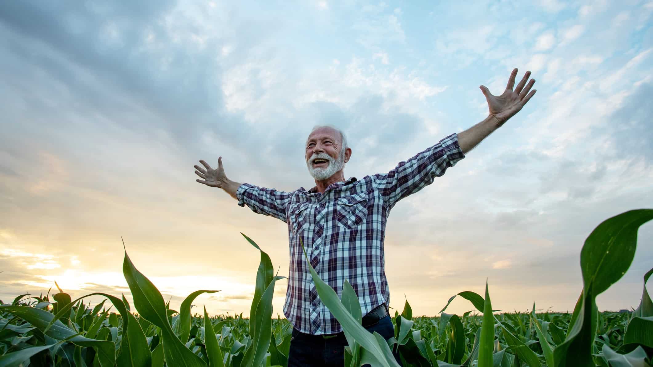 An older farmer stands arms outstretched in a field with a big smile on their face.