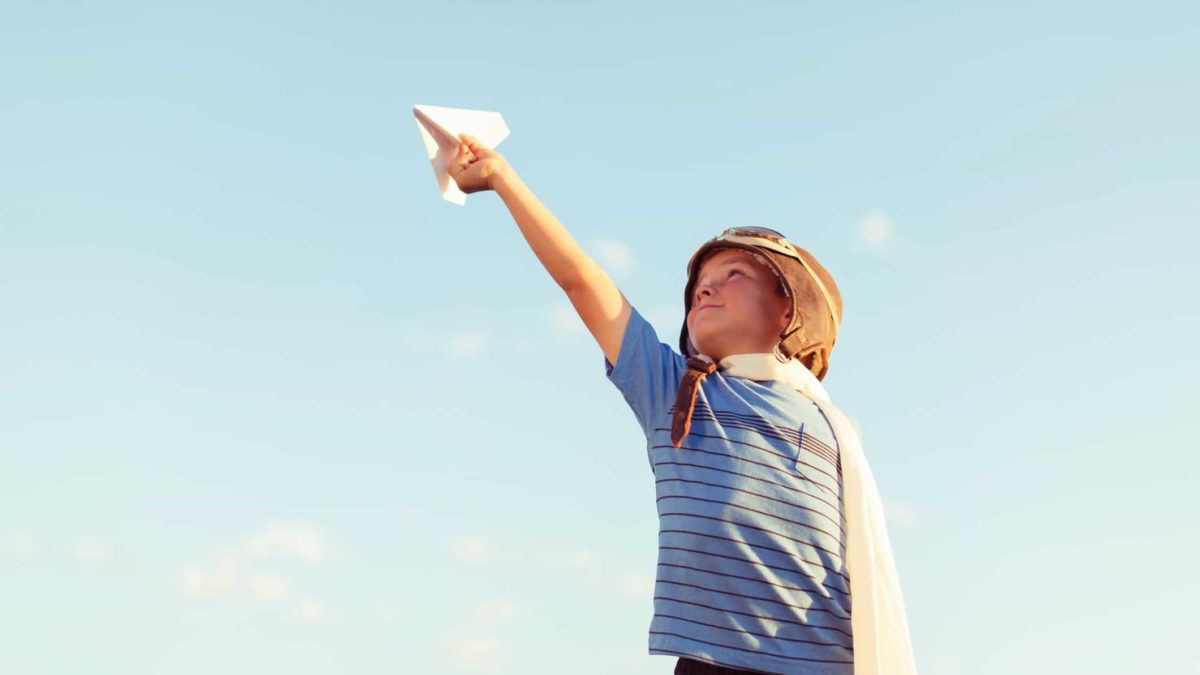 A kid wearing a pilot helmet holds a paper plane up to the sky.
