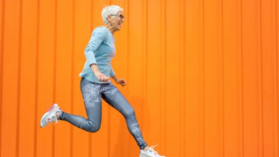 A fit older woman leaps in the air in front of a bright orange wall.