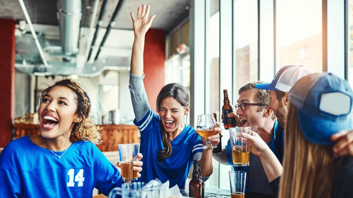 A group of friends watch the game at the pub whilst enjoying a few drinks, one girl has her hand up cheering.