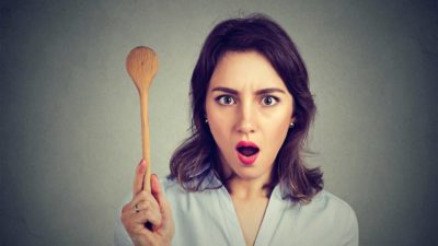 A woman holds a wooden spoon in her hand with a shocked look.