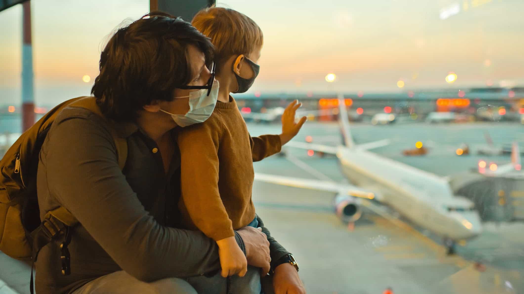 a father and his son wear masks and gaze out the window of an airport lounge onto planes on the tarmac below with an orange sunset glow in the background as they wonder whether Virgin Australia will relist on the ASX and become an ASX travel share again