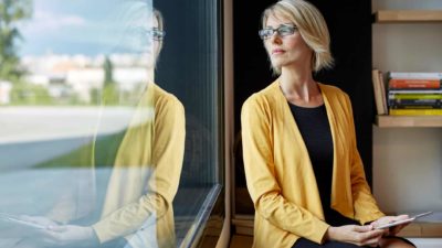 a woman wearing the black and yellow corporate colours of a leading bank gazes out the window in thought as she holds a tablet in her hands.