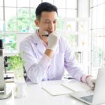 a biomedical researcher sits at his desk with his hand on his chin, thinking and giving a small smile with a microscope next to him and an array of test tubes and beackers behind him on shelves in a well-lit bright office.
