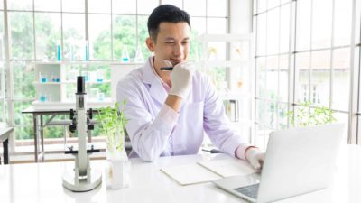 a biomedical researcher sits at his desk with his hand on his chin, thinking and giving a small smile with a microscope next to him and an array of test tubes and beackers behind him on shelves in a well-lit bright office.