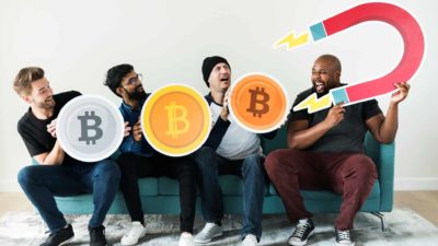 Three men sit in a row holding giant bitcoins while the fourth wields a huge magnet