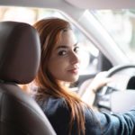 A woman looks over her shoulder towards the back seat while sitting at the wheel of a stationary car with a serious look on her face.